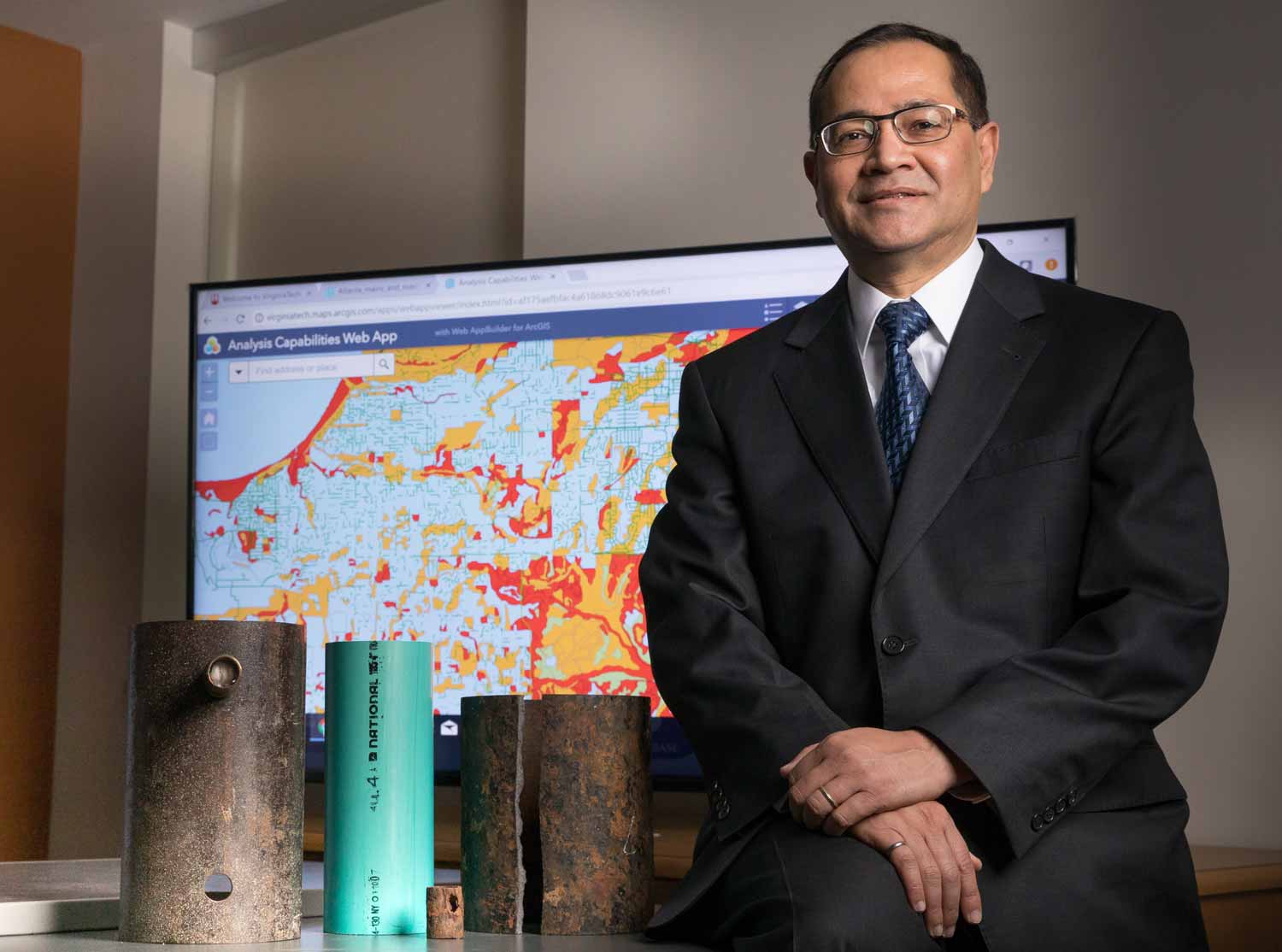 Sunhil Sinha poses with various rusty water pipe sections in front of a water map on a digital display.