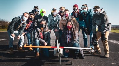 The Design Build Fly team poses for a group photo with a plane on a runway.