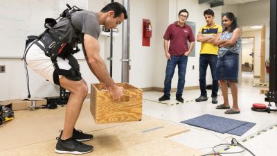 A Virginia Tech graduate student lifts a box while wearing an exoskeleton suite in a BEAM Engineering lab.