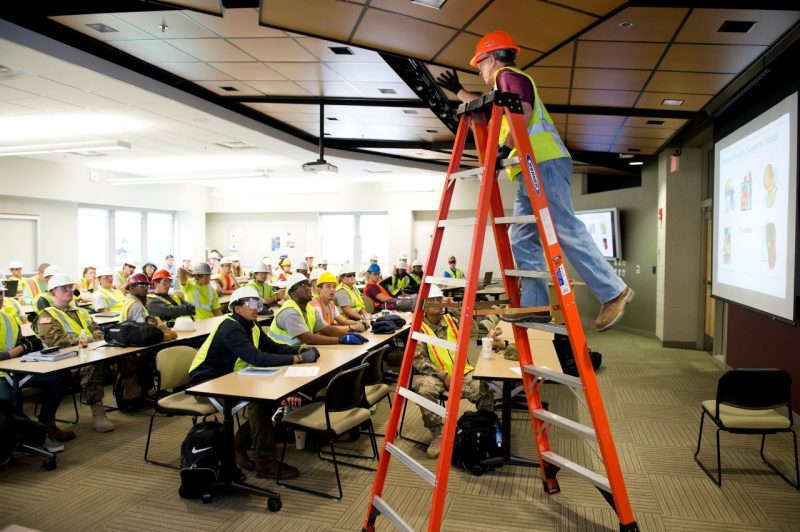 A man in a construction hat and vest stands on a ladder in the classroom