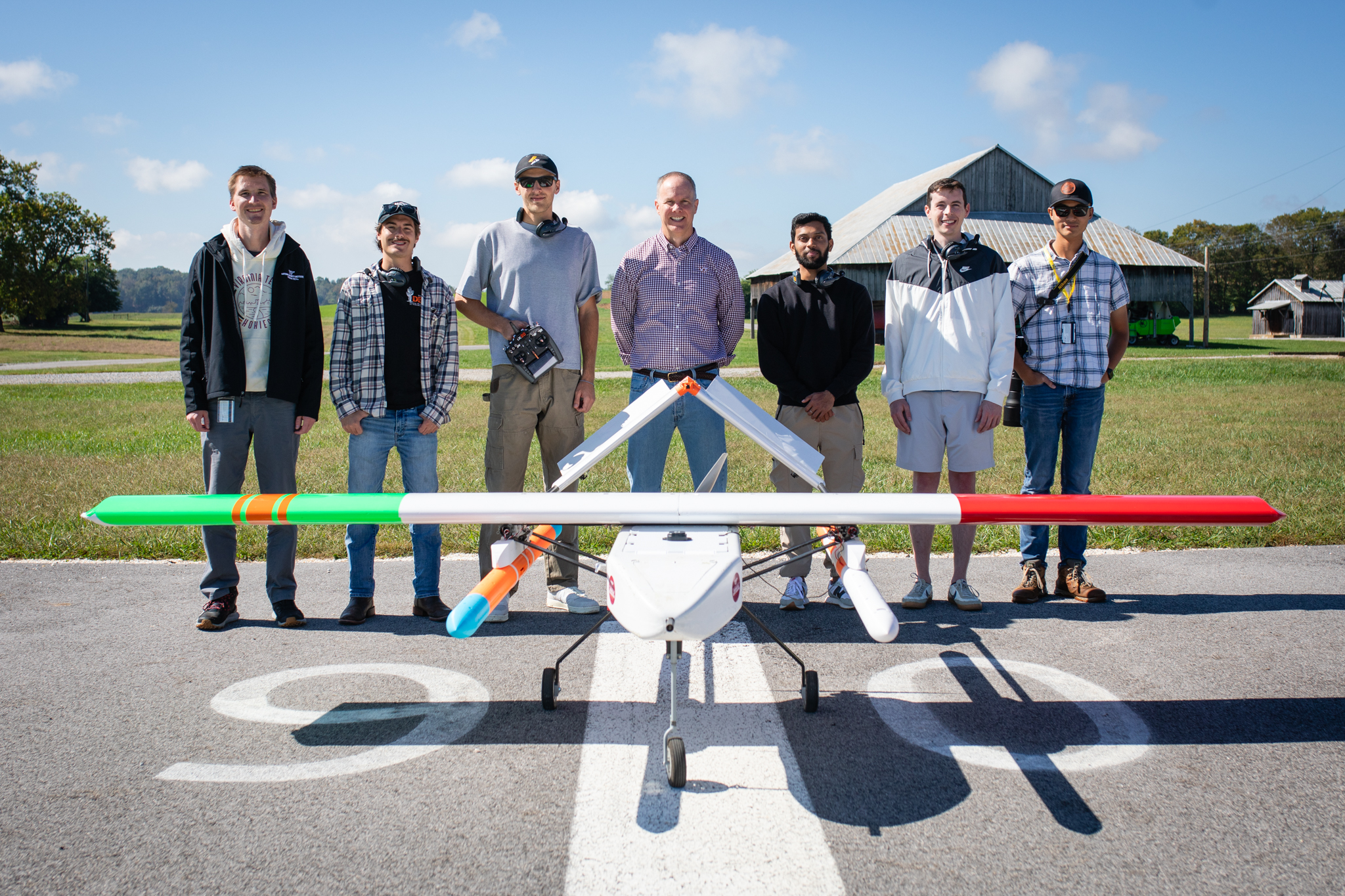 The team and faculty member pose for a photo behind a large weather drone on the end of a runway.