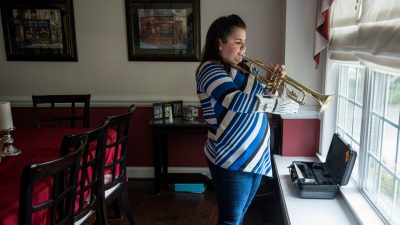 Josie Fraticelli, a young, 12-year-old girl, stands in front of a window in her home, playing a trumpet using a white prosthetic hand.