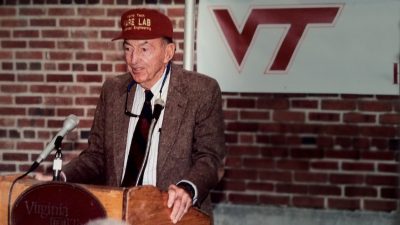 A photo of Joe Ware standing at a podium wearing a maroon hat embroidered with the words "WARE LAB" in uppercase, bold letters.
