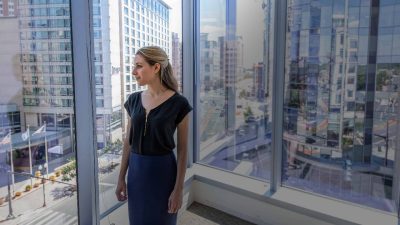 A woman stands in front of a window. Behind her stand tall buildings with large glass windows in a modern cityscape.