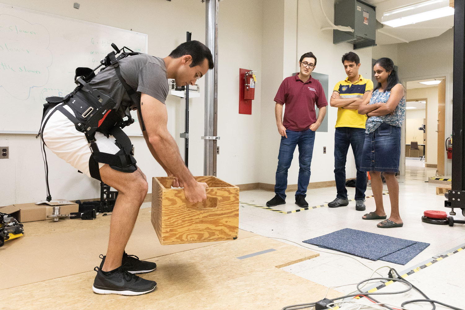 A group stands to the side and watches as a man wearing an exoskeleton lifts a wooden crate.