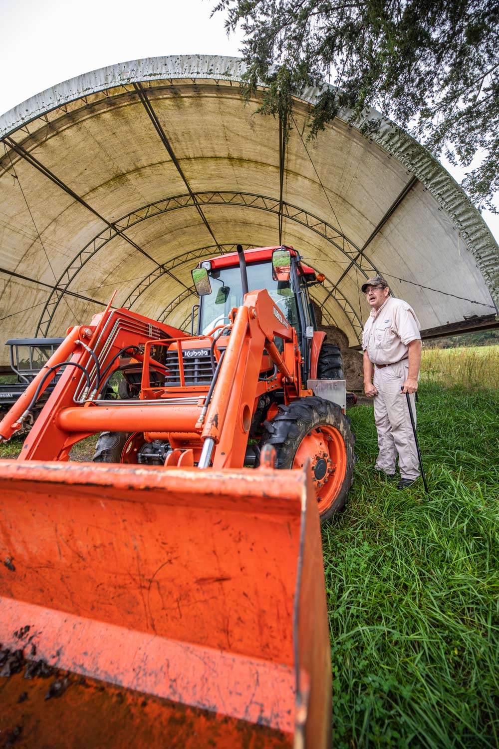 A man leans on a cane standing to the side of a bright orange tractor.