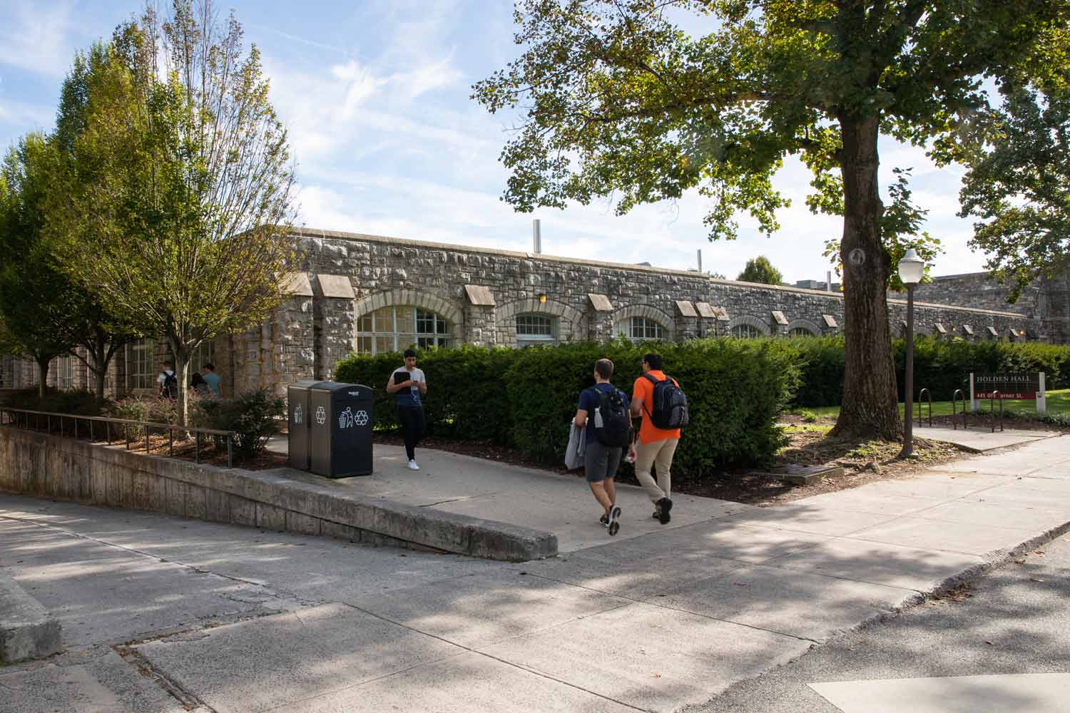 A few students walk on the sidewalk in front of an exterior of Holden Hall that faces Turner Street. It is one story tall and mostly covered from view with bushes.