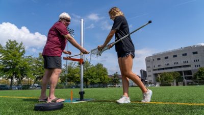 Two people use a lacrosse trainer on a lacrosse field.