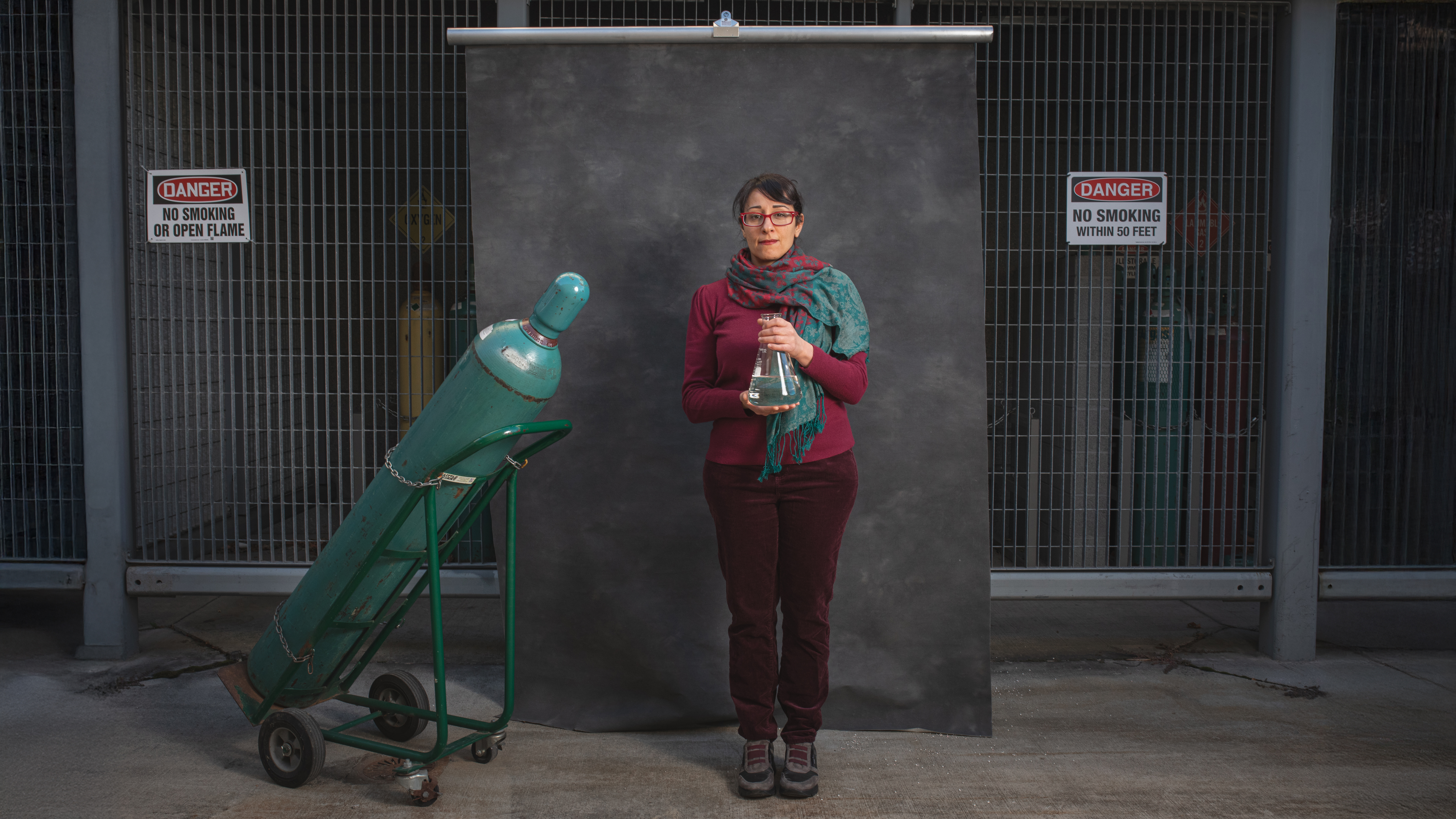 Sheima Khatib posing in front of a gas cylinder chamber holding a glass beaker.