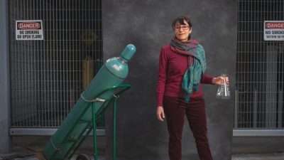 Sheima Khatib posing in front of a gas cylinder chamber holding a glass beaker.