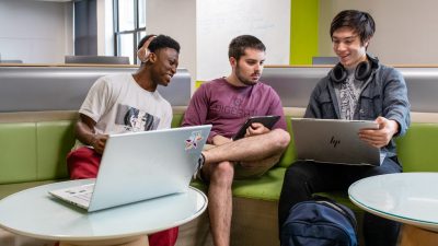 Three students work on their laptops together in a student lounge area.