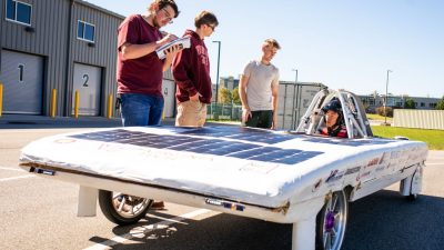 The SolarCar on the track with three students talking to the driver and taking notes.