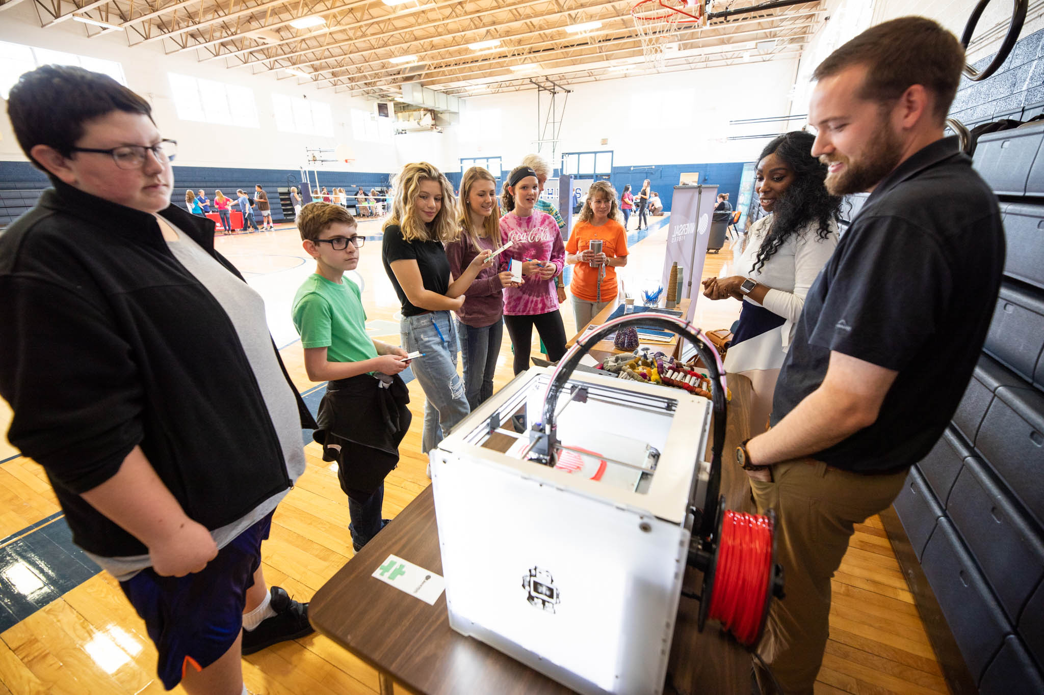 Young students watch a 3D printer in action on a folding table in a gymnasium.