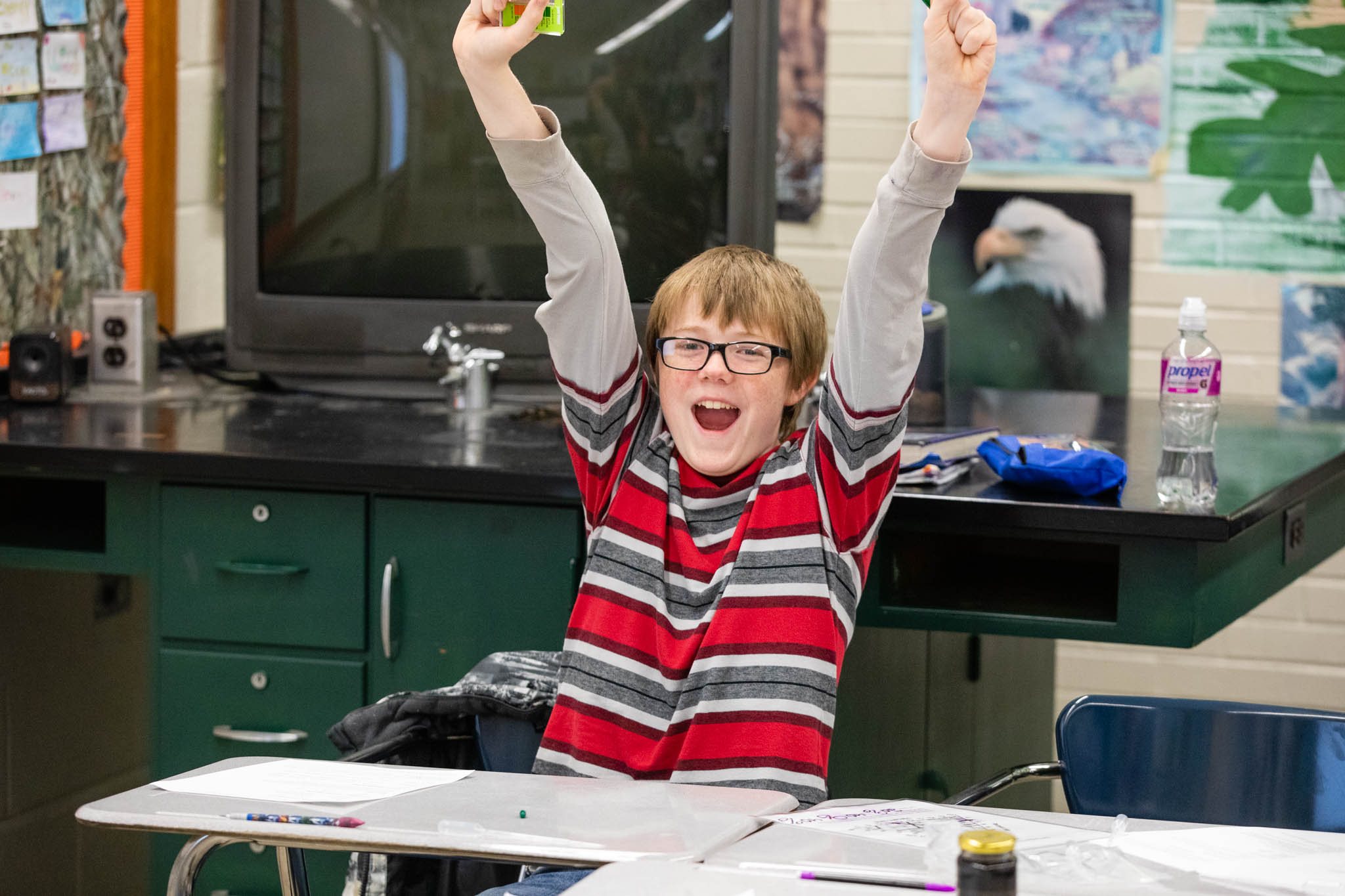 A young man throws his arms up in the air in celebration, in a classroom.