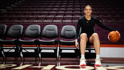 Taylor Geiman sitting in a row of chairs court-side bouncing a basketball.