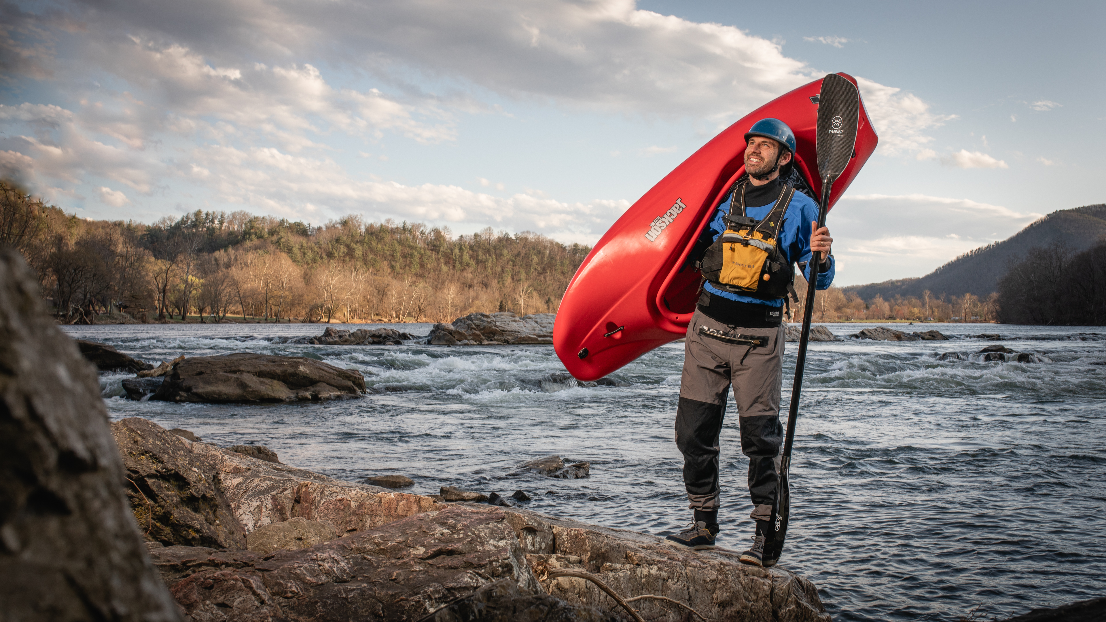 Tripp Shealy with a kayak over his shoulder, standing on the rocks next to a river.