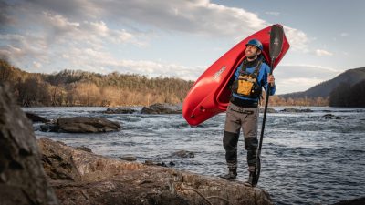 Tripp Shealy with a kayak over his shoulder, standing on the rocks next to a river.