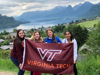 Group of four female-presenting individuals in Europe, mountains in the background, and holding up a Virginia Tech flag.