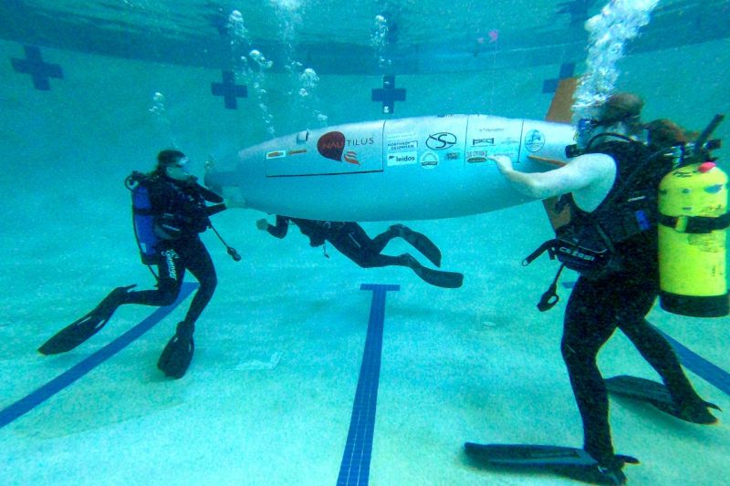 Three team members wearing SCUBA gear assist the sub under water in a pool.