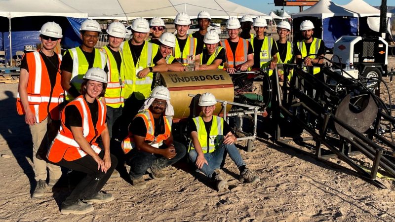 Diggeridoos team photo by their machine in a sandy location at competition. 