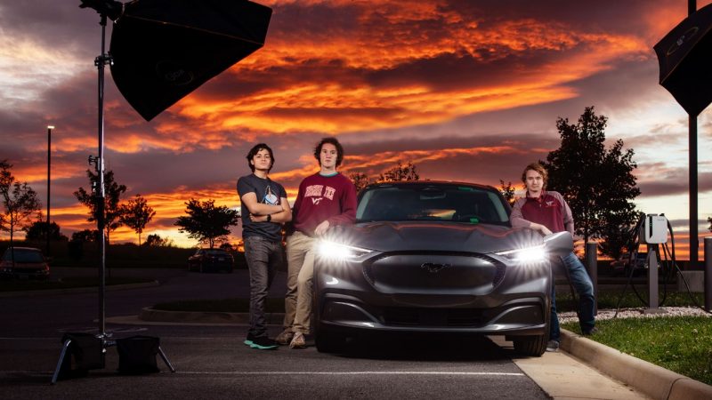 Three students posing by a Ford Mach-E car at sunset with photography lights.