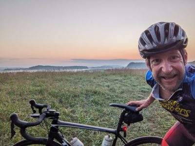 Aaron Goldstein takes a selfie with his bike in front of a sunset outdoor scene.