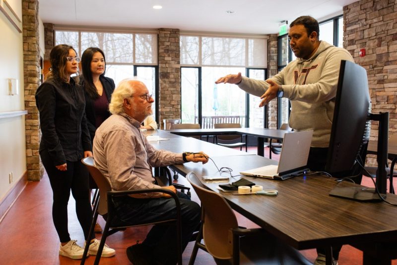Three researchers gather around a test subject at a table with a laptop, explaining the haptic device.