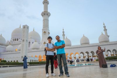 Two students stand smiling side-by-side, arms crossed, in front of the Grand Mosque in Abu Dhabi, which is tall, white, and topped with oval steeples.