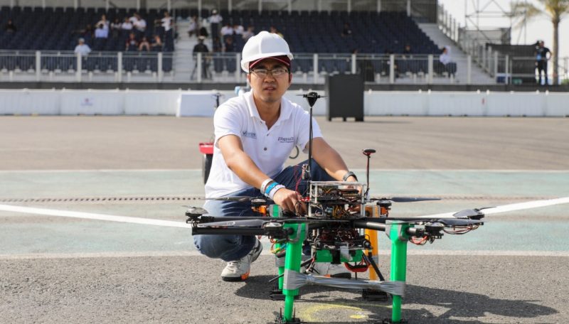 Student posing with a drone in a drone cage in front of stands where onlookers watch the competition.