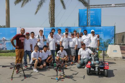 Students and professors gather for a group photo in front of a blue wall with their two drones and a ground vehicle positioned in front of them.