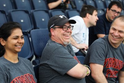 Three people wearing gray shirts that read "Virginia Tech" in orange and maroon laugh together while sitting in blue stands.