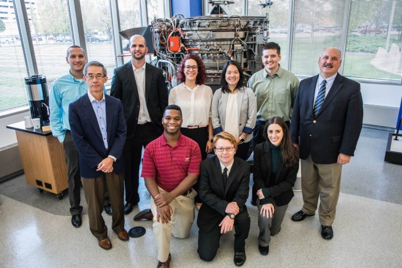 A group of students and researchers pose for a photo in front of a jet engine on the Rolls-Royce campus in Indianapolis.