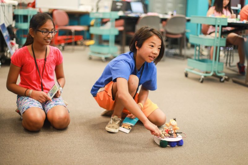 Two girls, about 10 years old, play on floor with a roving robot vacuum topped with stuffed animals in the Qualcomm Thinkabit Lab located in Falls Church, Virginia. 